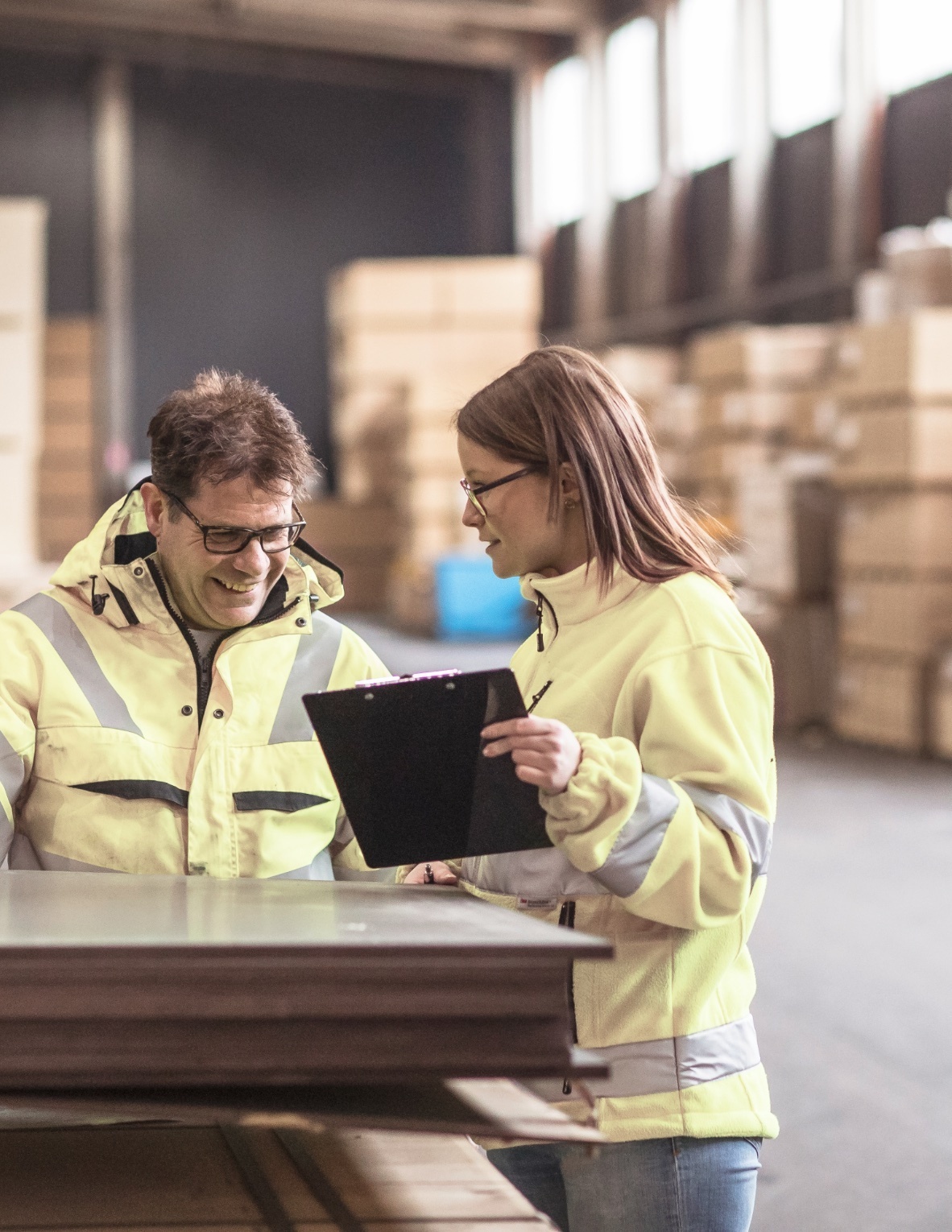 Två personer i reflexjackor står i ett lager och diskuterar. Den ena håller en mapp medan den andra ler och ser på dokumenten. Bakgrunden består av staplar med kartonger.