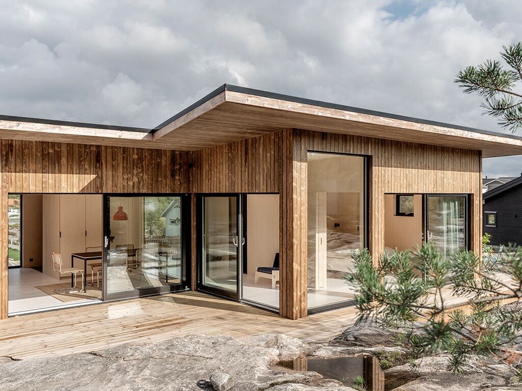 Modern enplanshus med träfasad och stora glaspartier, beläget på en klippig terräng. Utanför finns en träaltan som leder in till en ljus och öppen inredning. Molnig himmel i bakgrunden.
